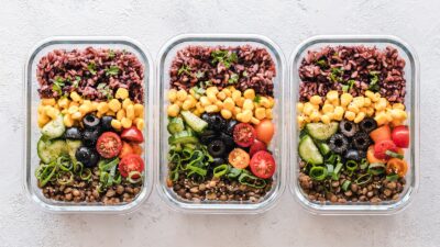 Glass meal-prep containers with roasted vegetables, whole grains, chickpeas, protein, greens, berries, nuts and lemon-herb dressing on a bright kitchen counter.