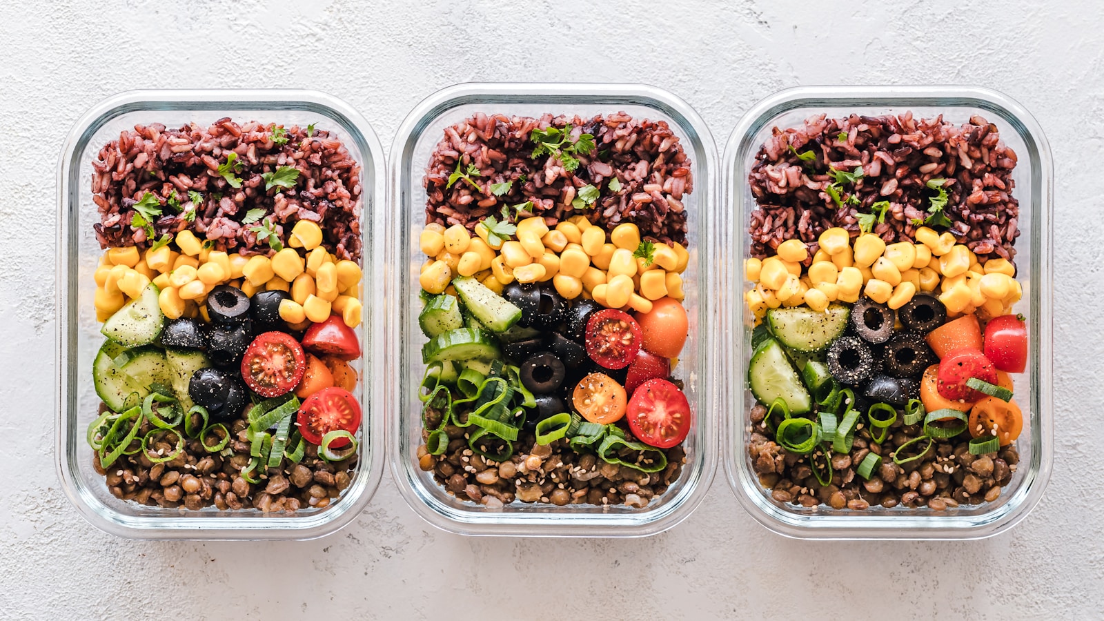 Glass meal-prep containers with roasted vegetables, whole grains, chickpeas, protein, greens, berries, nuts and lemon-herb dressing on a bright kitchen counter.